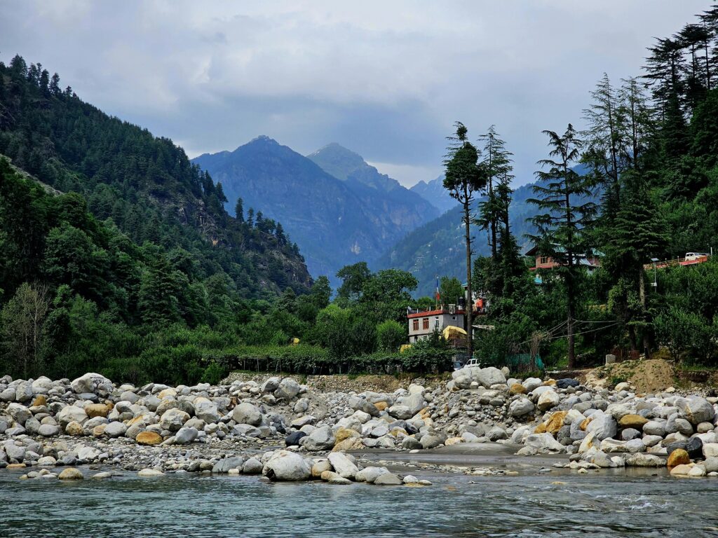 The Beas River flowing through a rocky riverbed, surrounded by lush green forests and majestic mountain ranges in Himachal Pradesh