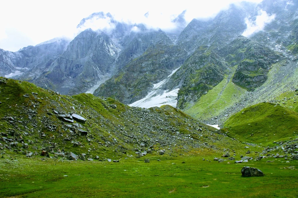 A vibrant green alpine meadow scattered with boulders, leading up to steep, cloud-covered Himalayan mountains with snow patches on the Beas Kund trek.
