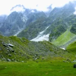 A vibrant green alpine meadow scattered with boulders, leading up to steep, cloud-covered Himalayan mountains with snow patches on the Beas Kund trek.