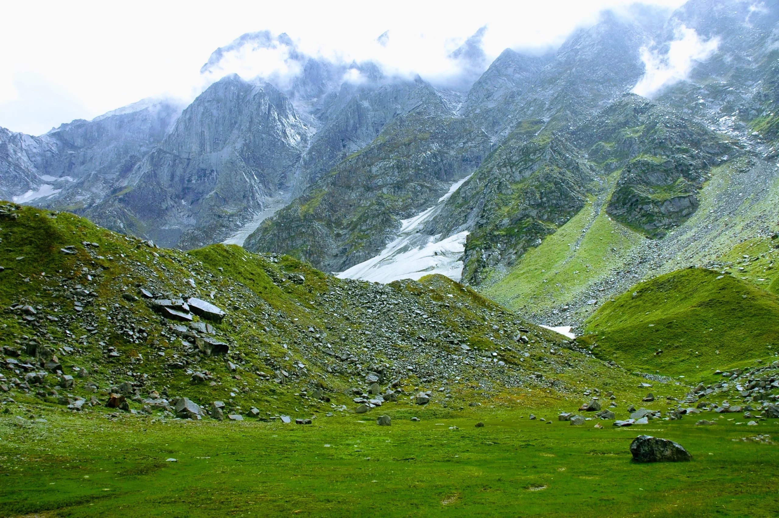 A vibrant green alpine meadow scattered with boulders, leading up to steep, cloud-covered Himalayan mountains with snow patches on the Beas Kund trek.