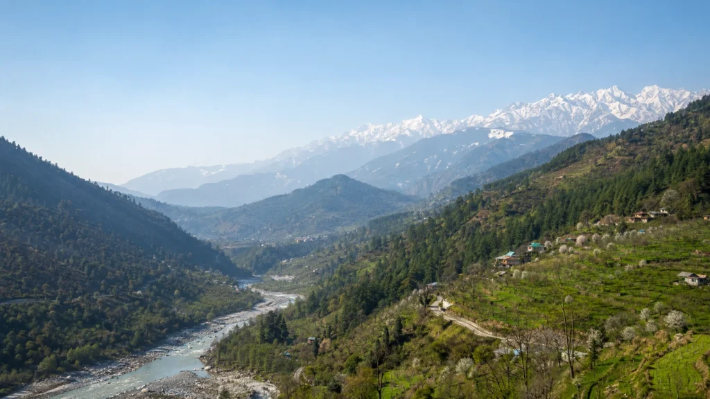 Himalayan valley with a river, forested slopes, terraced orchards, hill settlements and distant snow-covered peaks.